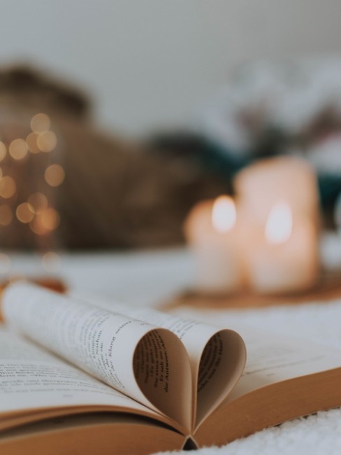 Libro abierto con páginas dobladas en forma de corazón, acompañado de velas encendidas al fondo sobre una superficie suave, creando un ambiente cálido y acogedor.